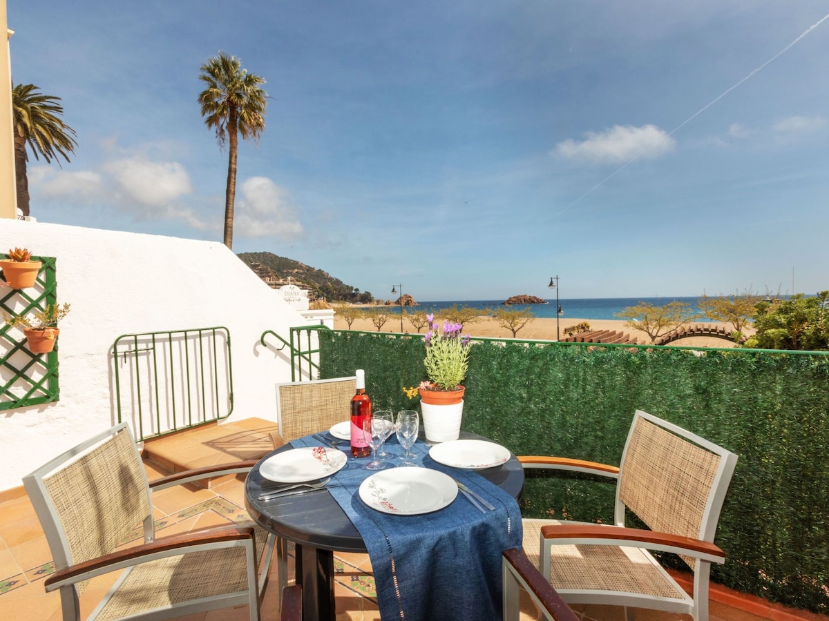 A spacious balcony features a round dining table set with plates and glasses. A potted plant adds a touch of greenery, while unobstructed views of the beach and ocean can be seen in the background. Sunlight brightens the outdoor space, complementing the coastal scenery.