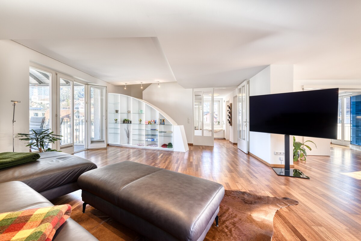 A modern living area features a large sectional couch with earthy tones, complemented by a stylish area rug. Expansive windows provide ample natural light and views of the surroundings. A flat-screen TV is positioned for easy viewing, while decorative shelving adds an element of sophistication.