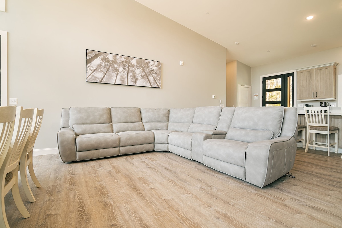 A spacious living area features a large sectional sofa in light grey fabric, complemented by light wooden flooring. A dining table with white chairs is visible in the background. A wall-mounted artwork depicts palm trees, enhancing the room's relaxed ambiance.
