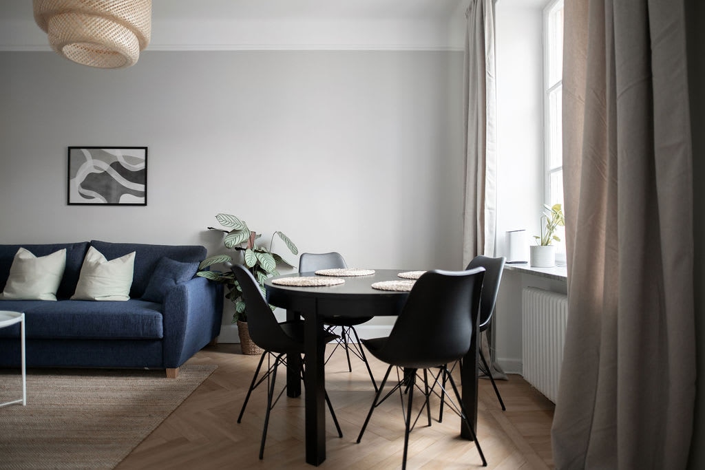 A bright living area features a dark blue sofa adorned with light-colored cushions, complemented by a round black dining table surrounded by four black chairs. Natural light floods the room through large windows, illuminating the smooth wooden floor and indoor plants nearby.