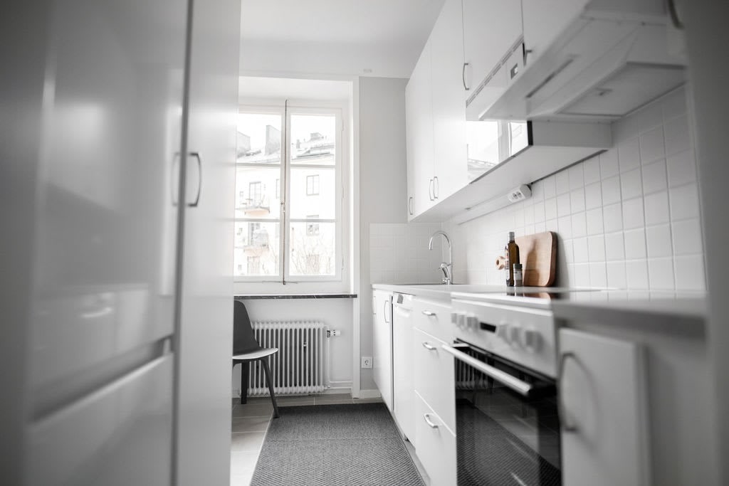 A bright kitchen features modern, white cabinetry with ample storage and a sleek countertop. Natural light enters through a window above the sink, while essential appliances such as an oven and refrigerator are positioned conveniently. A small dining chair rests against the wall.