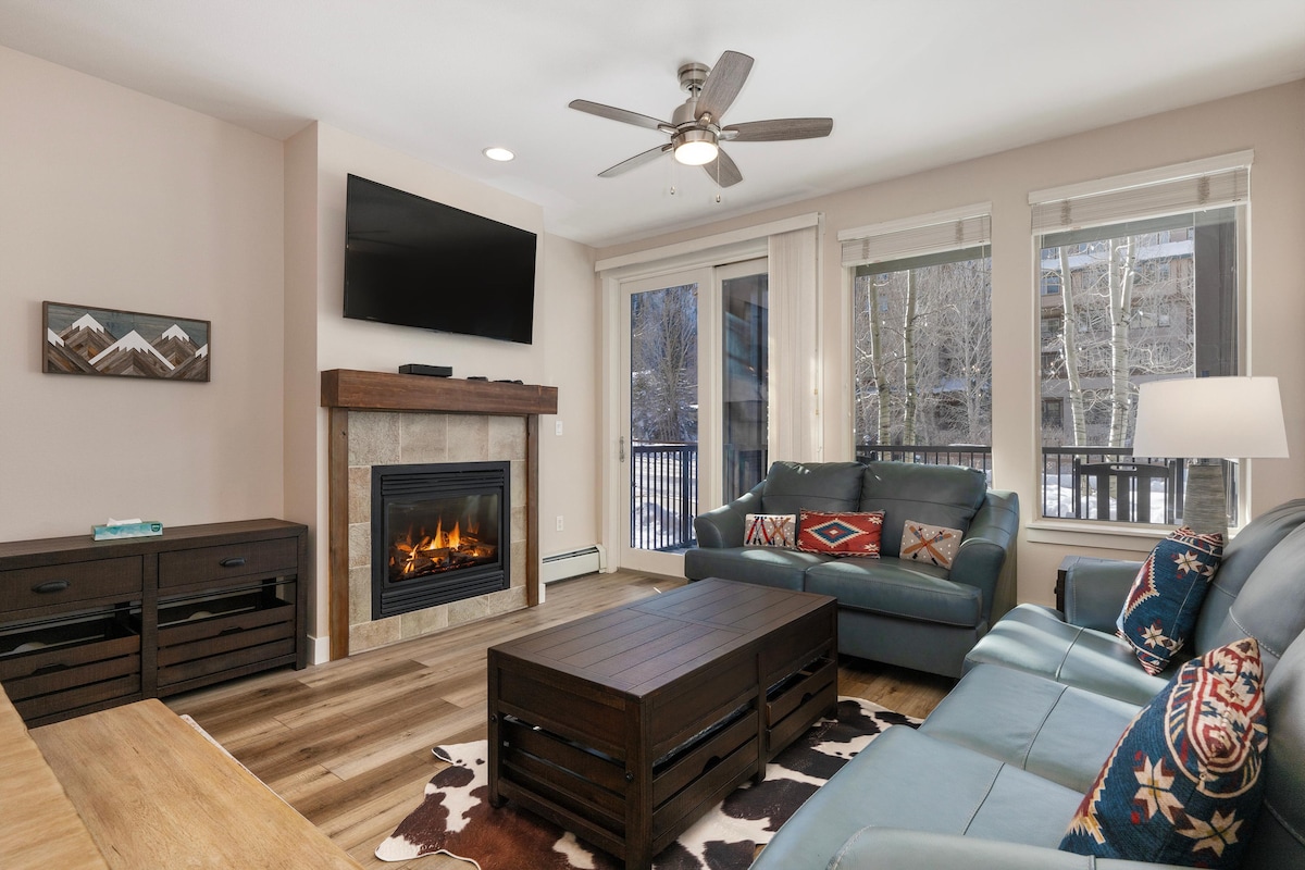 A spacious living area is highlighted by a cozy gas fireplace and a mounted flat-screen TV. Comfortable leather sofas, adorned with patterned cushions, surround a rustic coffee table. Large sliding glass doors provide natural light and access to a balcony with views of the surrounding nature.