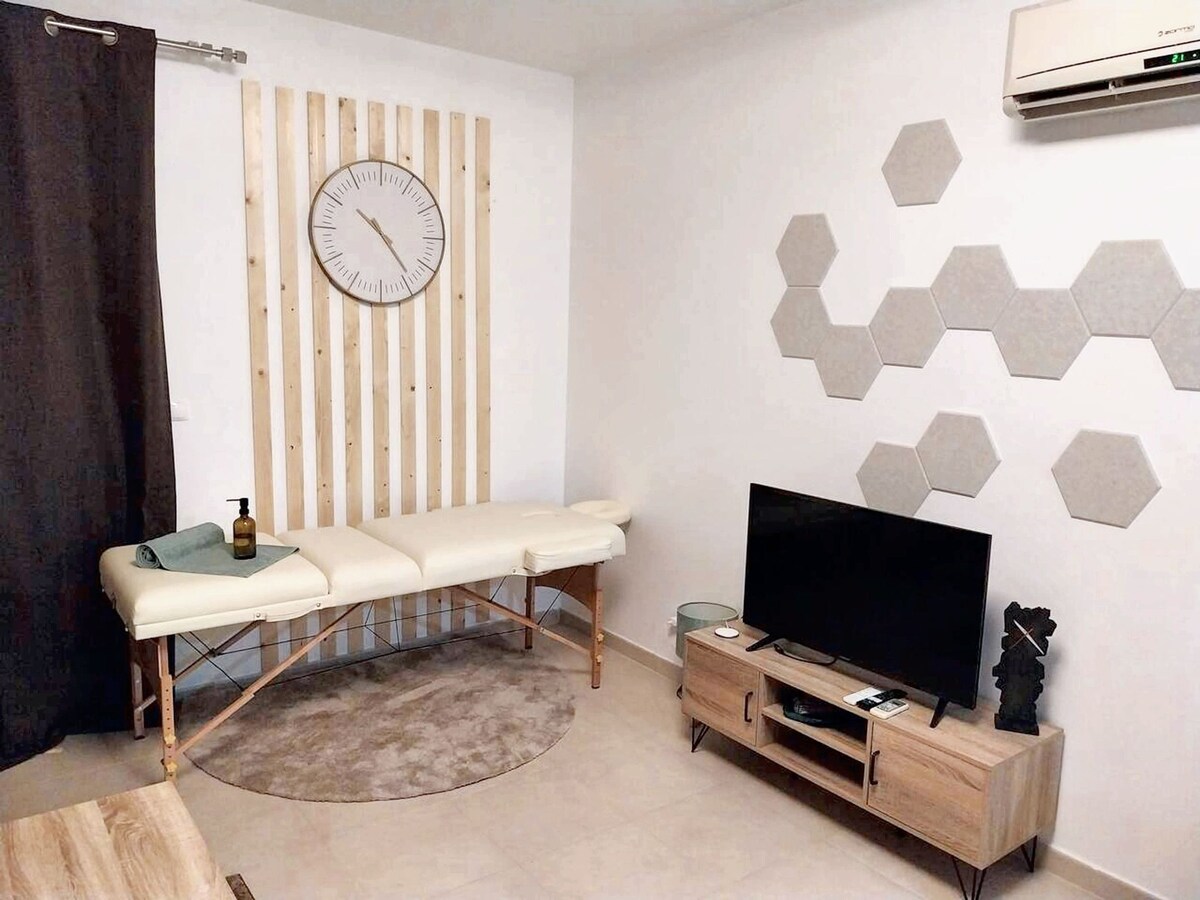 A soothing wellness space features a massage table with a rolled towel, complemented by a wall clock above. Hexagonal wall panels add texture, while a television sits on a wooden entertainment unit. Natural light enhances the calming ambiance of this area.