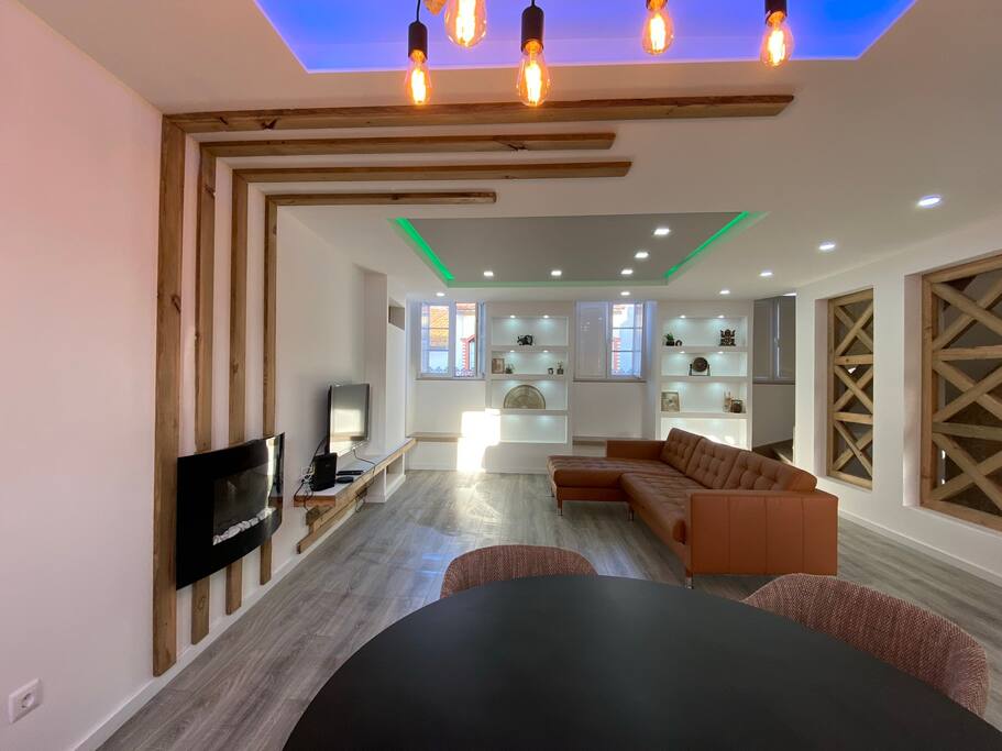 A modern living area is displayed, featuring a comfortable brown sectional sofa and a sleek black dining table. Built-in shelves with decorative items are arranged in the background. Natural light enters through the large windows, while stylish lighting fixtures illuminate the space.
