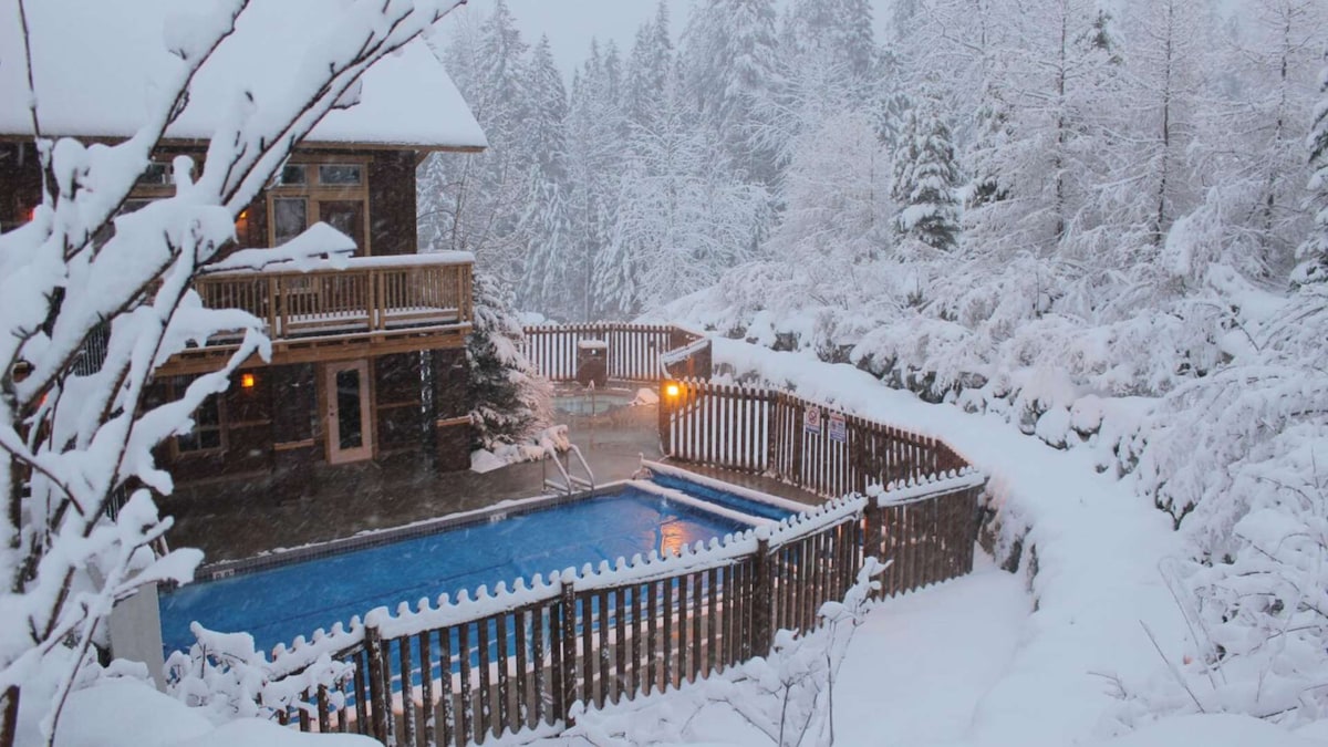 An outdoor heated pool is framed by a wooden fence, surrounded by freshly fallen snow and pine trees. The building features multiple balconies, with warm light visible through the windows, creating a cozy ambiance against the winter landscape.