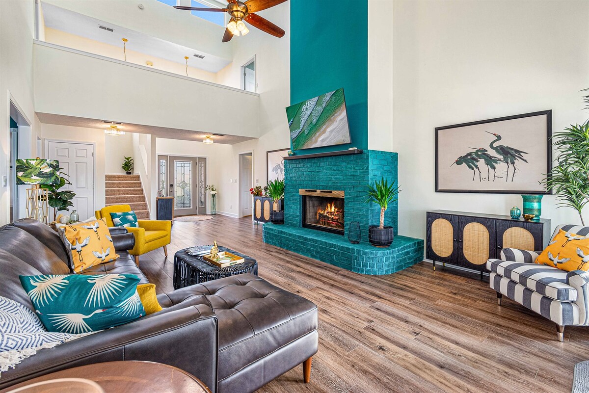 A spacious living area features comfortable seating arranged around a coffee table. A striking green accent wall with a fireplace is complemented by a large television above. Natural light pours in through the windows, enhancing the inviting atmosphere of the room.
