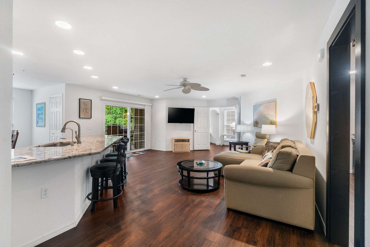 An open living area is presented with a contemporary sofa and armchairs, complemented by a mounted smart TV. A breakfast bar with stools lines the kitchen, featuring glossy countertops and stainless steel appliances. Natural light fills the space through glass doors leading to an outdoor area.