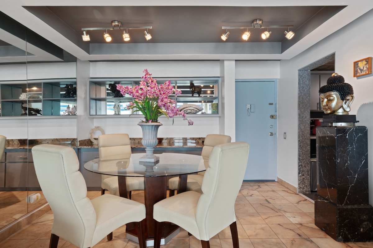 A modern dining area is showcased, featuring a round glass table surrounded by four ivory chairs. A vibrant floral arrangement sits at the center, adding a touch of color. Reflections from the mirror wall enhance the sense of space, while overhead lighting illuminates the area.