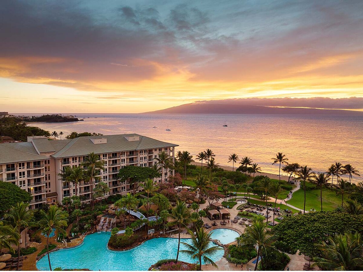 A panoramic view captures the resort's lush grounds with palm trees lining the pool area. The horizon showcases a vibrant sunset over the ocean, with distant boats visible. The accommodations are positioned to highlight the tranquil atmosphere of the beachfront location.