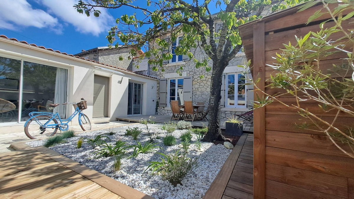 A charming exterior view of the house is presented, featuring a landscaped garden with white stones and greenery. Comfortable wooden lounge chairs are arranged on a patio area. A bicycle is parked nearby, and the house's windows reflect the blue sky above.