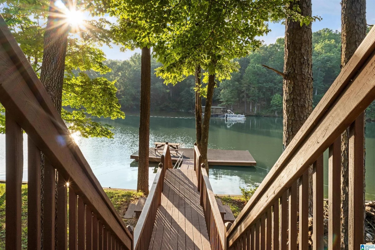 A wooden walkway leads down to a dock on a serene lake, framed by tall trees. Sunlight filters through the leaves, creating dappled light on the water's surface. The tranquil scene emphasizes the peaceful lakeside environment.