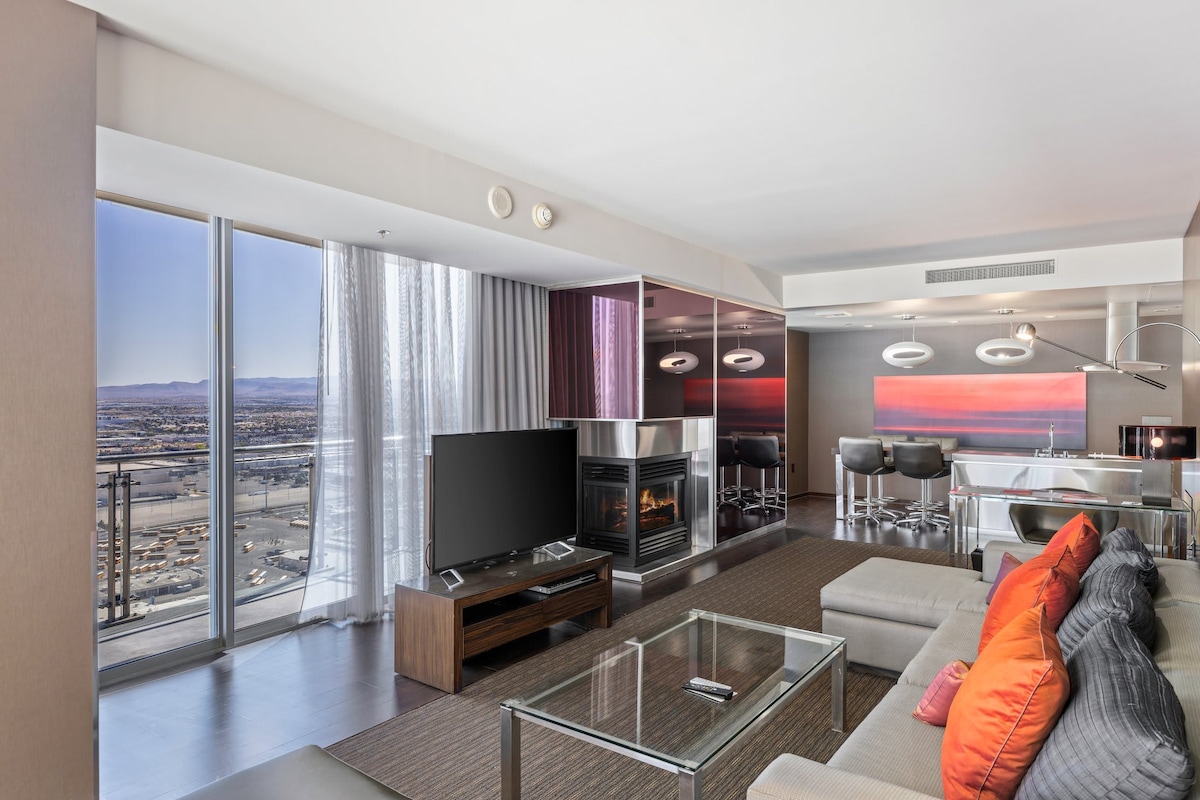 A stylish living area features large windows showcasing expansive mountain and city views. A modern fireplace adds warmth, while a flat-screen TV and a glass coffee table provide contemporary elements. Comfortable seating is adorned with vibrant cushions, enhancing the room's inviting atmosphere.