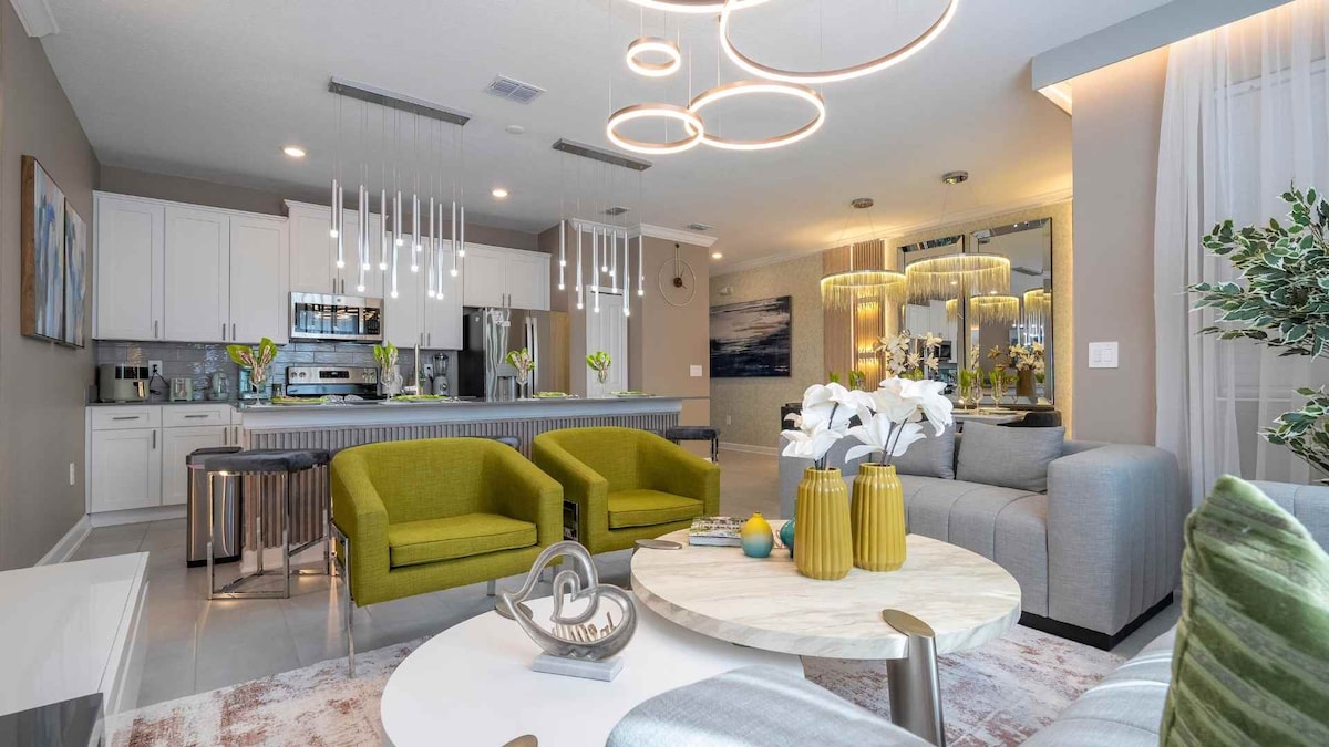 The spacious living area features contemporary furnishings, including two vibrant green chairs and a modern sofa, arranged around a round coffee table. Sleek kitchen appliances are visible in the background, along with illuminated pendant lights hanging above, creating a welcoming atmosphere.