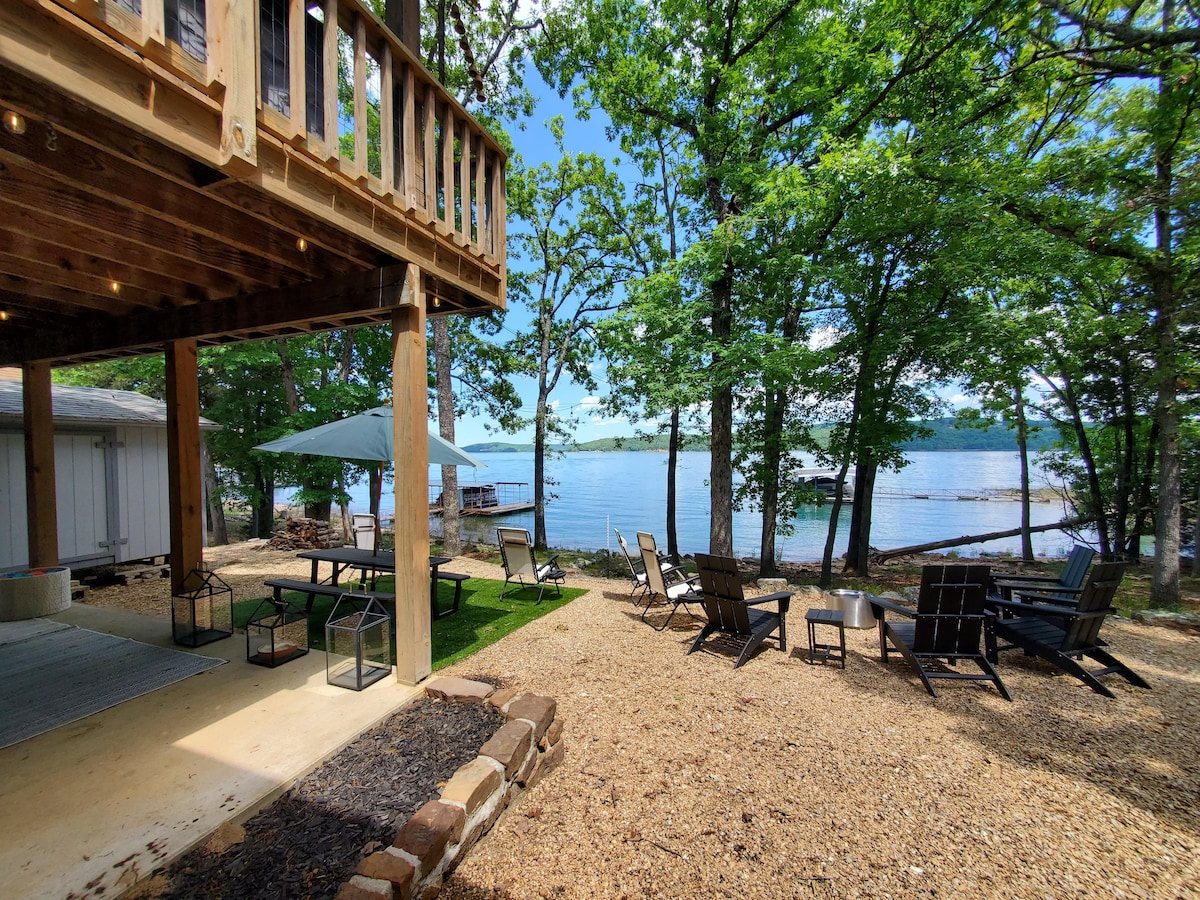 A serene lakeside scene is presented, with a covered patio overlooking the water. Several outdoor seating arrangements feature lounging chairs and a table. Tall trees provide shade, while gravel pathways guide to the water's edge, creating a peaceful setting for relaxation.