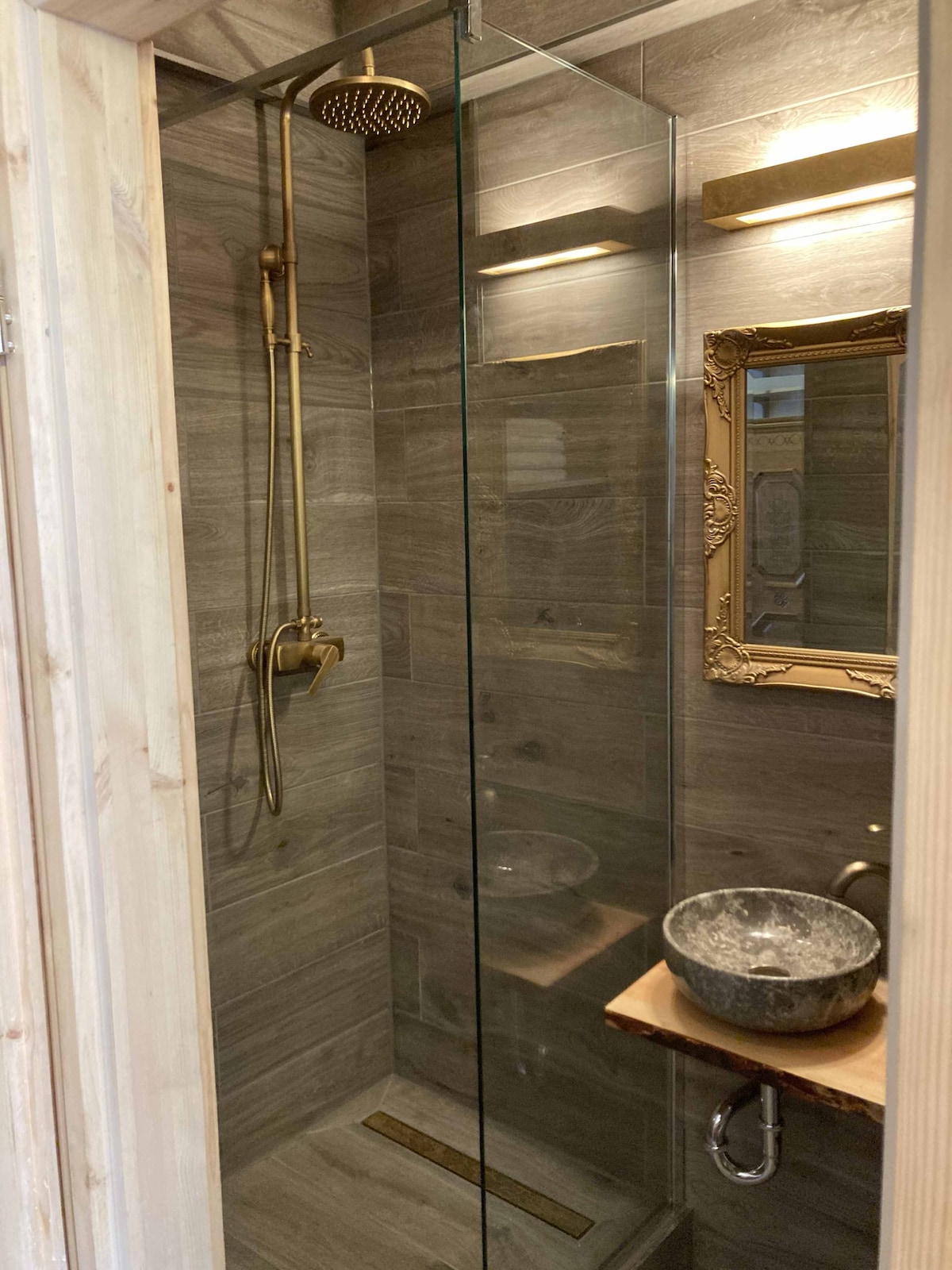 A modern shower area is presented, featuring a glass enclosure and elegant brass fixtures. The wooden paneling complements the stone sink on a natural wood shelf, while a decorative mirror with an ornate frame adds a touch of sophistication.