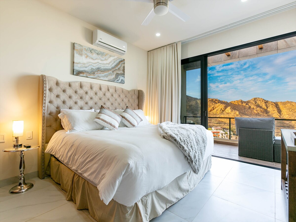 A spacious bedroom features a tufted headboard and a large bed with neutral bedding. A cozy throw is draped across the bed. Natural light floods the room through expansive glass doors, revealing a view of the mountainous landscape outside.