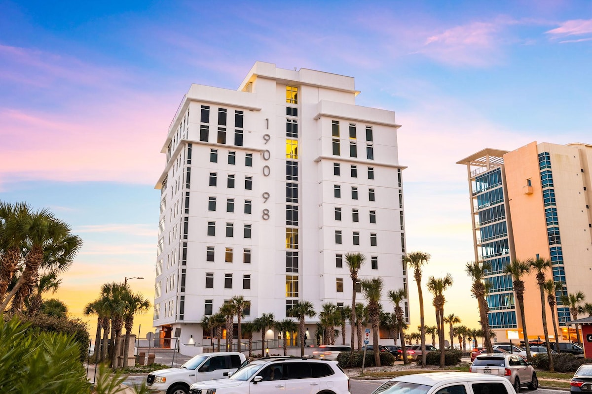 The modern condominium building is prominently displayed with its large, white façade and the number 1900 prominently featured. Surrounding palm trees add tropical elements, while vehicles are parked in front. A colorful sunset creates a backdrop of soft pastel hues in the sky.