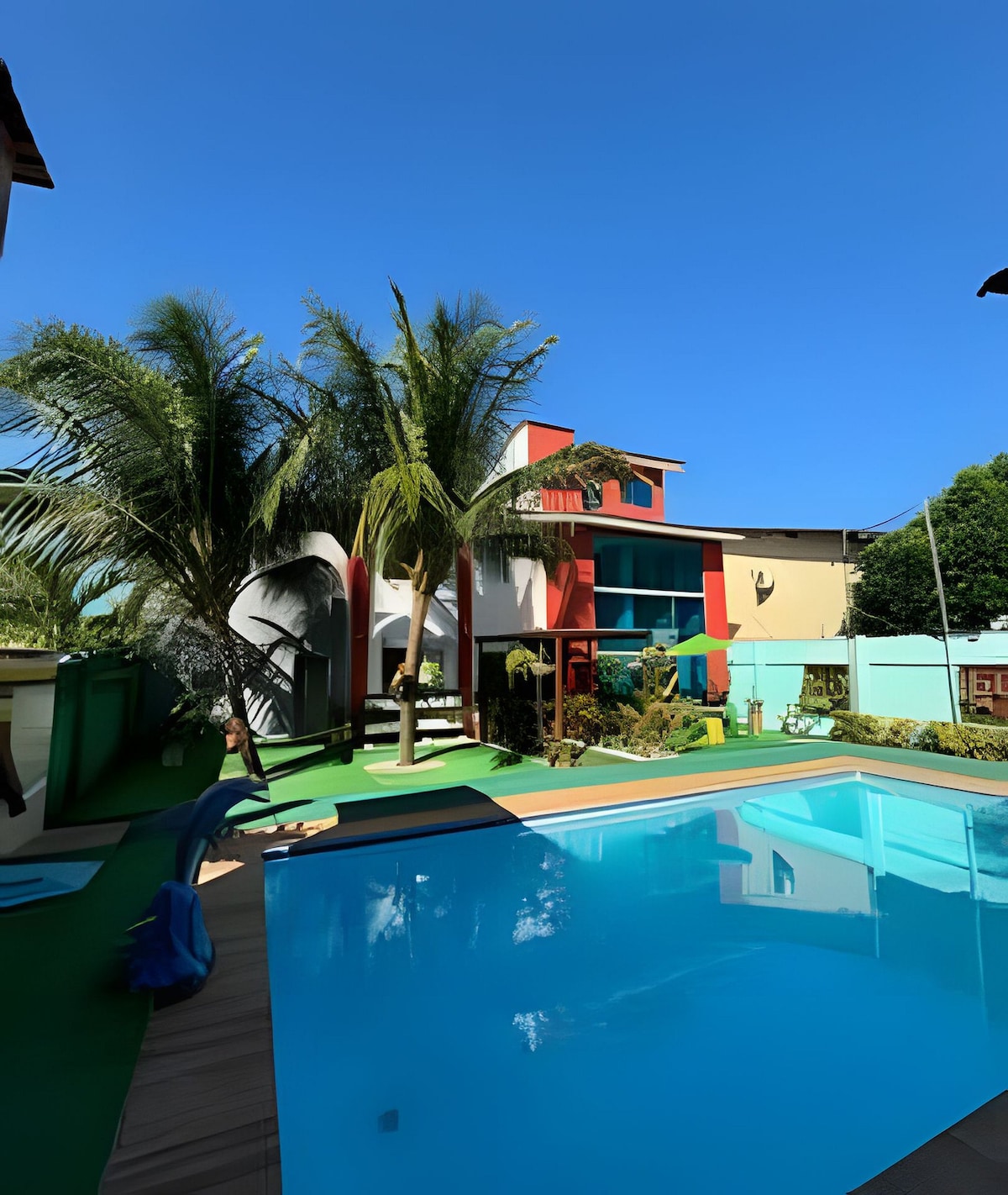 A vibrant exterior scene captures a well-maintained pool surrounded by lush greenery and palm trees. The brightly colored two-story house stands in the background under a clear blue sky, reflecting a welcoming atmosphere and easy access to outdoor leisure.