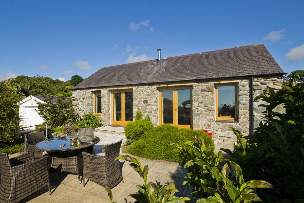 A stone cottage exterior is shown, featuring large glass panelled doors that open to a spacious patio area. A round dining table with four wicker chairs is set in front, surrounded by lush greenery. Clear blue skies complement the tranquil setting.