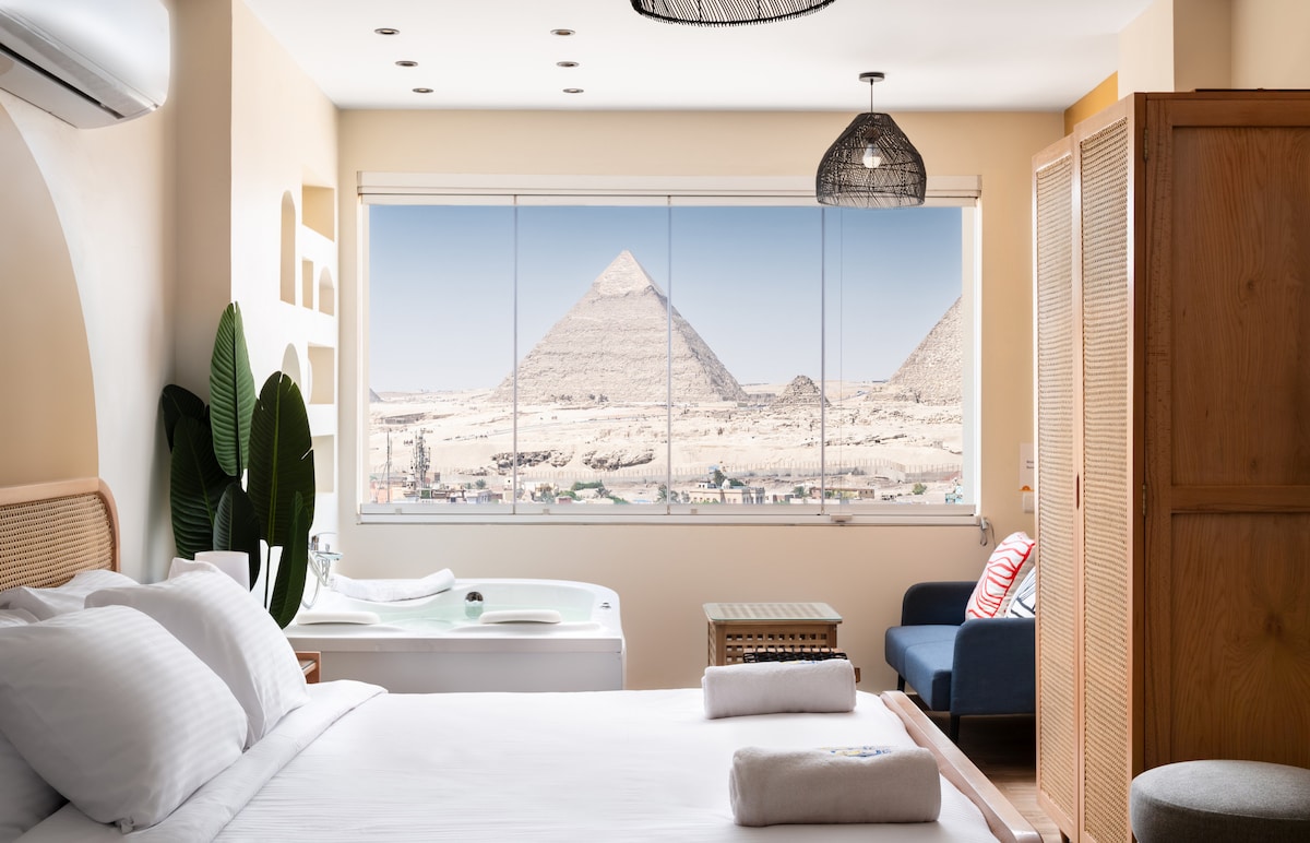 A spacious studio offers a panoramic view of the Pyramids of Giza through large windows. A comfortable king-size bed is positioned in front, with soft linens neatly arranged. A sparkling jacuzzi is visible alongside, and a cozy seating area is set by the window.
