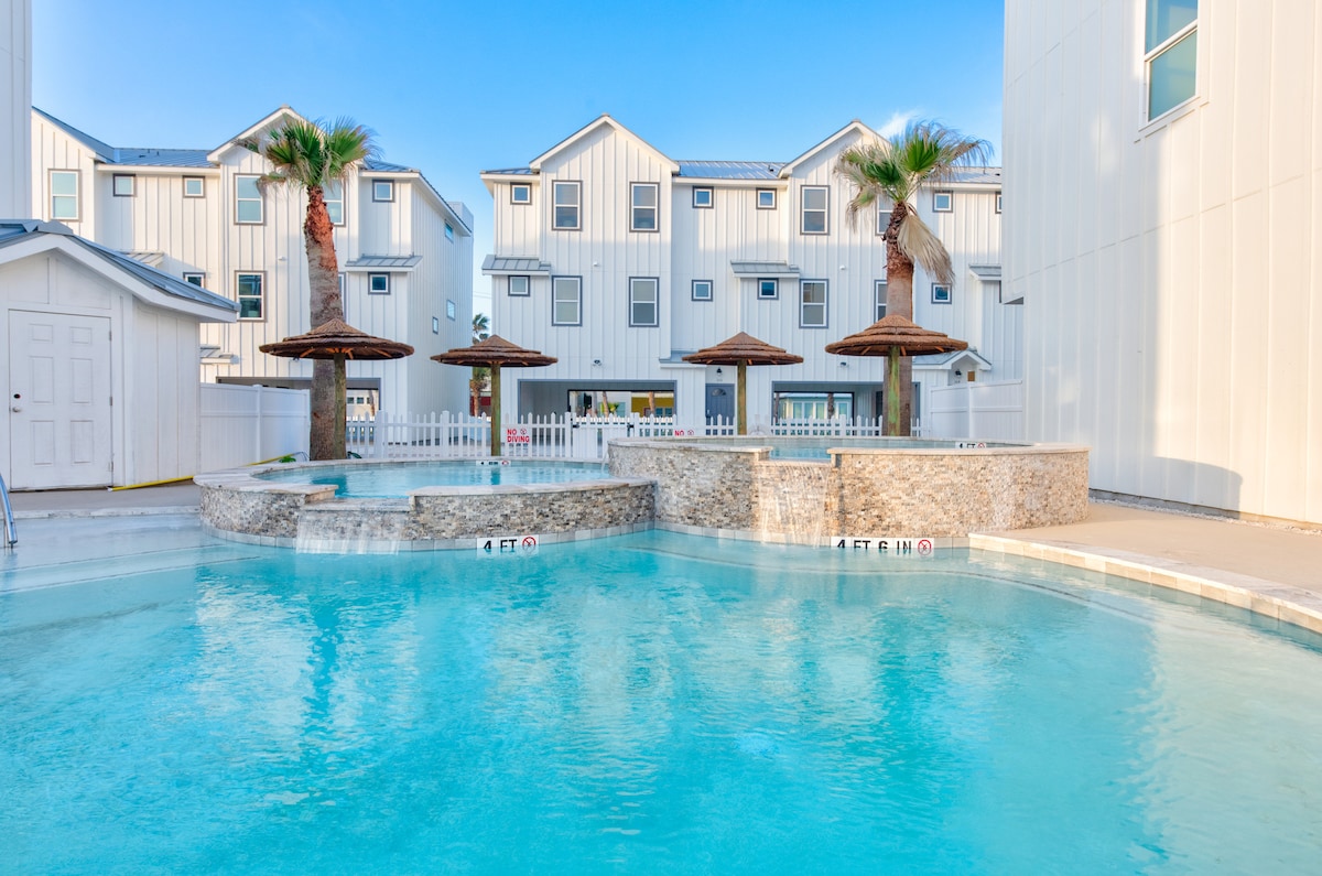 A sparkling pool area is showcased, featuring a large water body surrounded by palm trees and shaded loungers. The backdrop consists of white townhomes, enhancing the coastal ambiance, while a safety sign indicates depth levels for guest awareness.