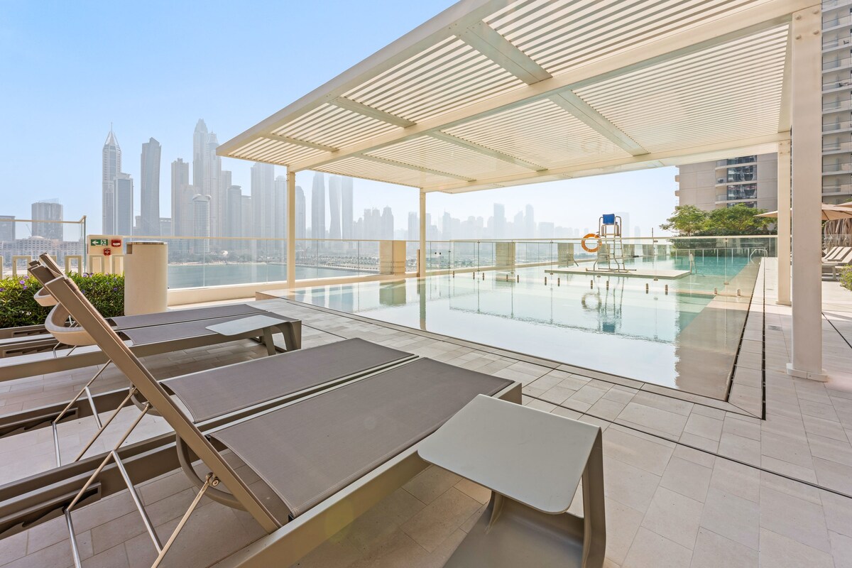 An outdoor pool area is presented with sleek sun loungers, offering views of the Dubai skyline. The pool reflects the surrounding cityscape, while a shaded pergola provides a comfortable space for relaxation. Clear skies enhance the bright and open atmosphere of the setting.