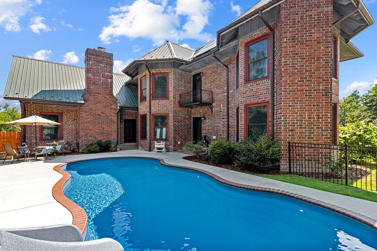 The backyard showcases a spacious inground pool surrounded by a brick patio, complemented by lush landscaping. The two-story house features a combination of brick and wood siding with multiple windows, enhancing the natural light. Lounge chairs are placed nearby, creating a relaxing atmosphere.