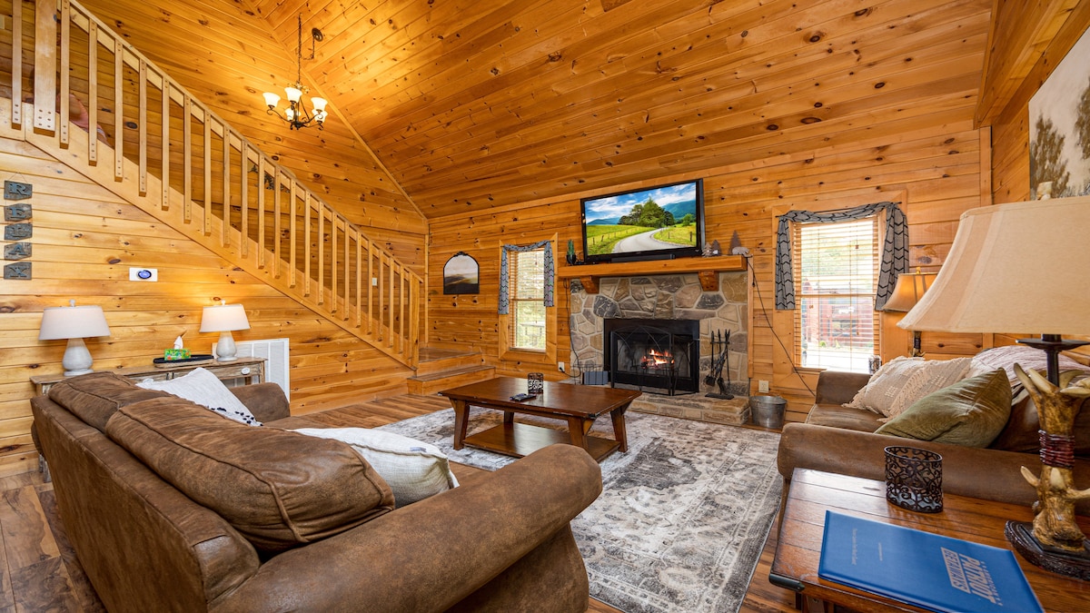 A warm and inviting living area features wood-paneled walls and a high ceiling, creating an open atmosphere. A leather sofa and a fabric couch are positioned around a wooden coffee table, while a stone fireplace adds a focal point. A television is mounted above the fireplace.