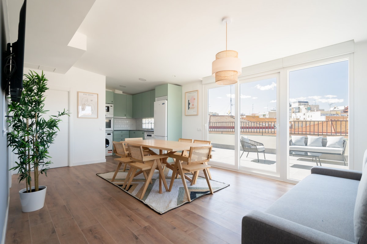 A bright dining area features a wooden table surrounded by six chairs, complemented by a light fixture overhead. Large windows allow for ample natural light and showcase views of the terrace. A sofa sits nearby, enhancing the open, airy feel of the space.