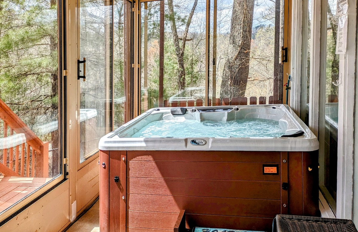 An inviting hot tub is positioned on an enclosed porch, surrounded by large glass windows that provide views of the wooded area outside. The bubbling water reflects the natural light streaming in, creating a serene atmosphere for relaxation.