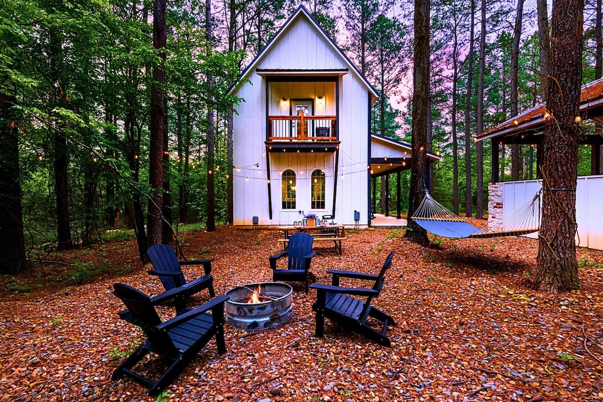 An outdoor seating area features five black Adirondack chairs arranged around a fire pit, set on a carpet of leaves. A hammock hangs between trees, and string lights illuminate the space. A two-story cabin, surrounded by woods, stands in the background.