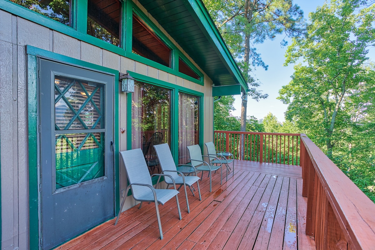 A spacious outdoor deck is seen, featuring five chairs positioned for relaxation. The deck overlooks lush greenery, providing a serene atmosphere. Large glass doors lead into the cabin, complementing the inviting outdoor space.