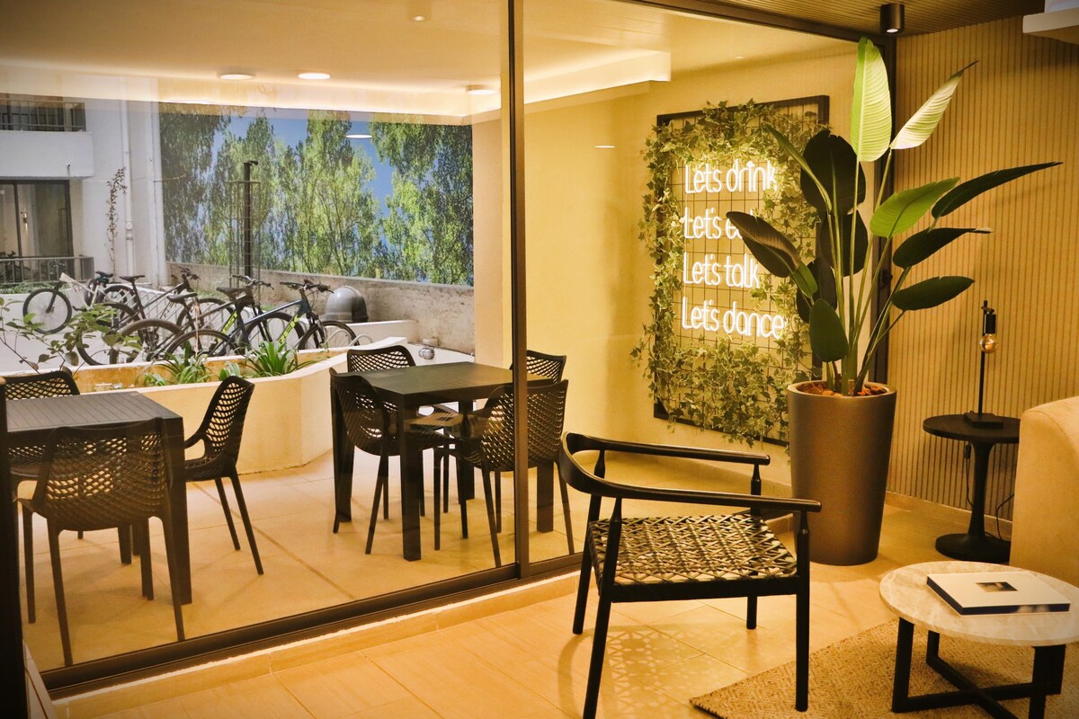An inviting terrace area is visible, featuring multiple outdoor seating arrangements with stylish black chairs and tables. A green plant stands in a large pot, and bicycles are parked in the background. Natural light floods the space through large glass doors.