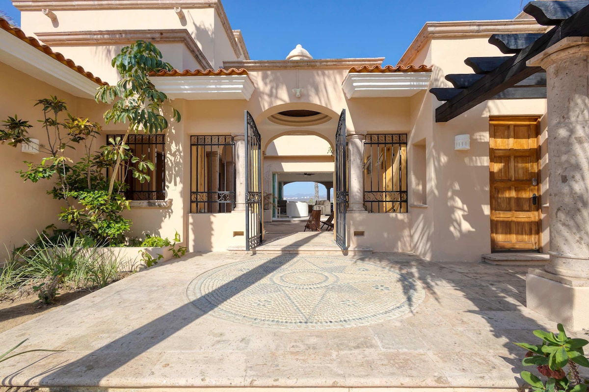 An open-air courtyard entrance features a decorative stone mosaic and is framed by arched doorways. Lush greenery and structured plants line the path, while the inviting wooden door and wrought iron accents add to the architectural charm of the villa.