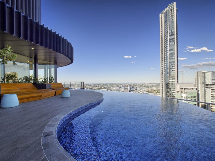 An infinity pool is seen on a rooftop, providing panoramic views of the surrounding cityscape. The pool's edge blends seamlessly into the horizon, enhancing the feeling of openness. Modern seating is placed nearby, complemented by potted greenery, creating a refreshing environment.