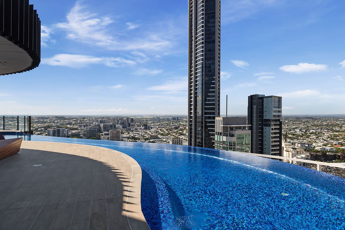 The rooftop pool offers a stunning view of the city skyline, with a sleek design and a shimmering blue surface. Surrounding areas feature modern buildings against a clear sky, enhancing the urban atmosphere. The pool edge appears to blend with the horizon, inviting relaxation.