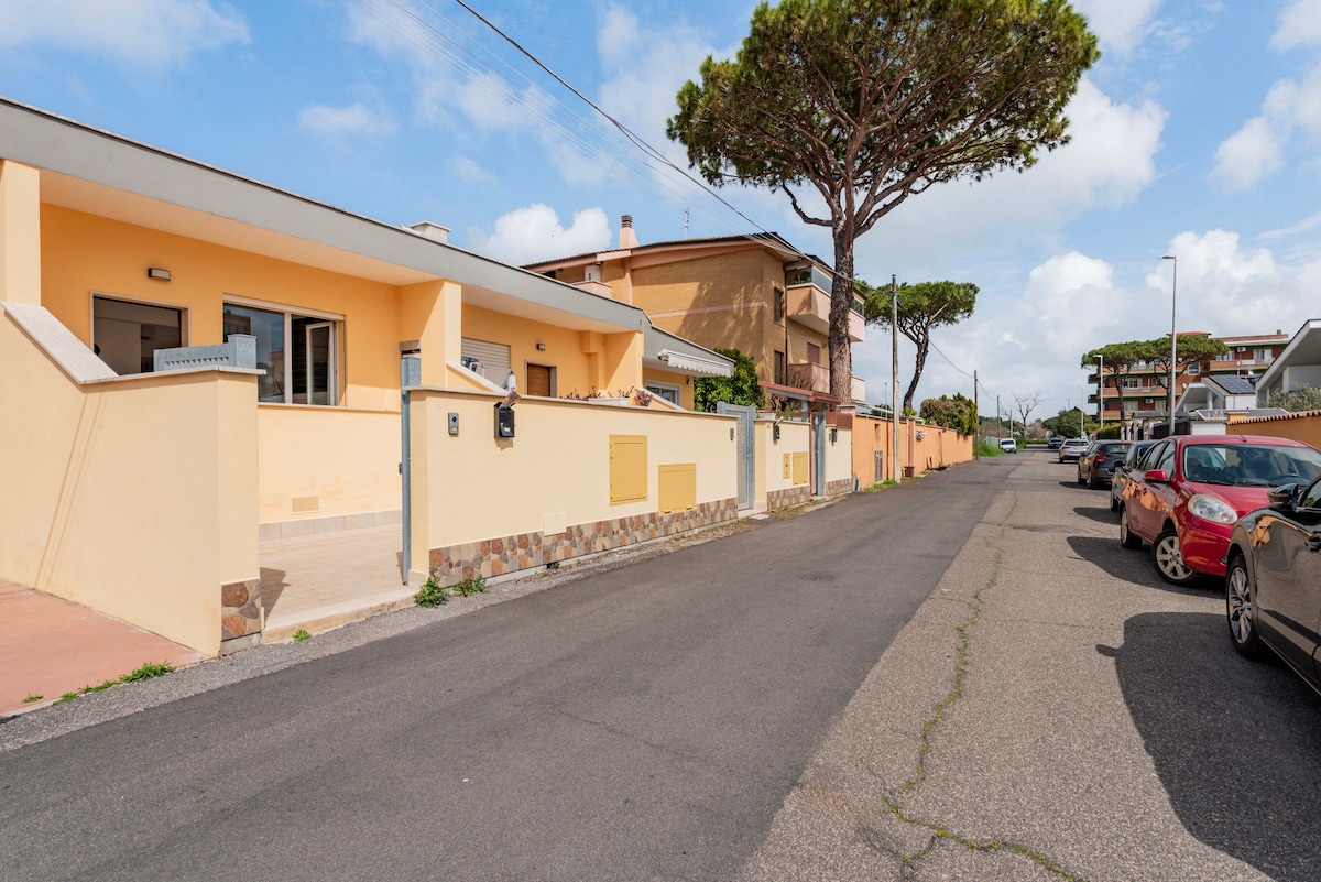 A quiet residential street is depicted, lined with parked cars and flanked by low-rise buildings. In the foreground, a well-maintained property features a warm yellow exterior and an accessible entrance. Lush trees provide shade, contributing to the pleasant atmosphere of the area.