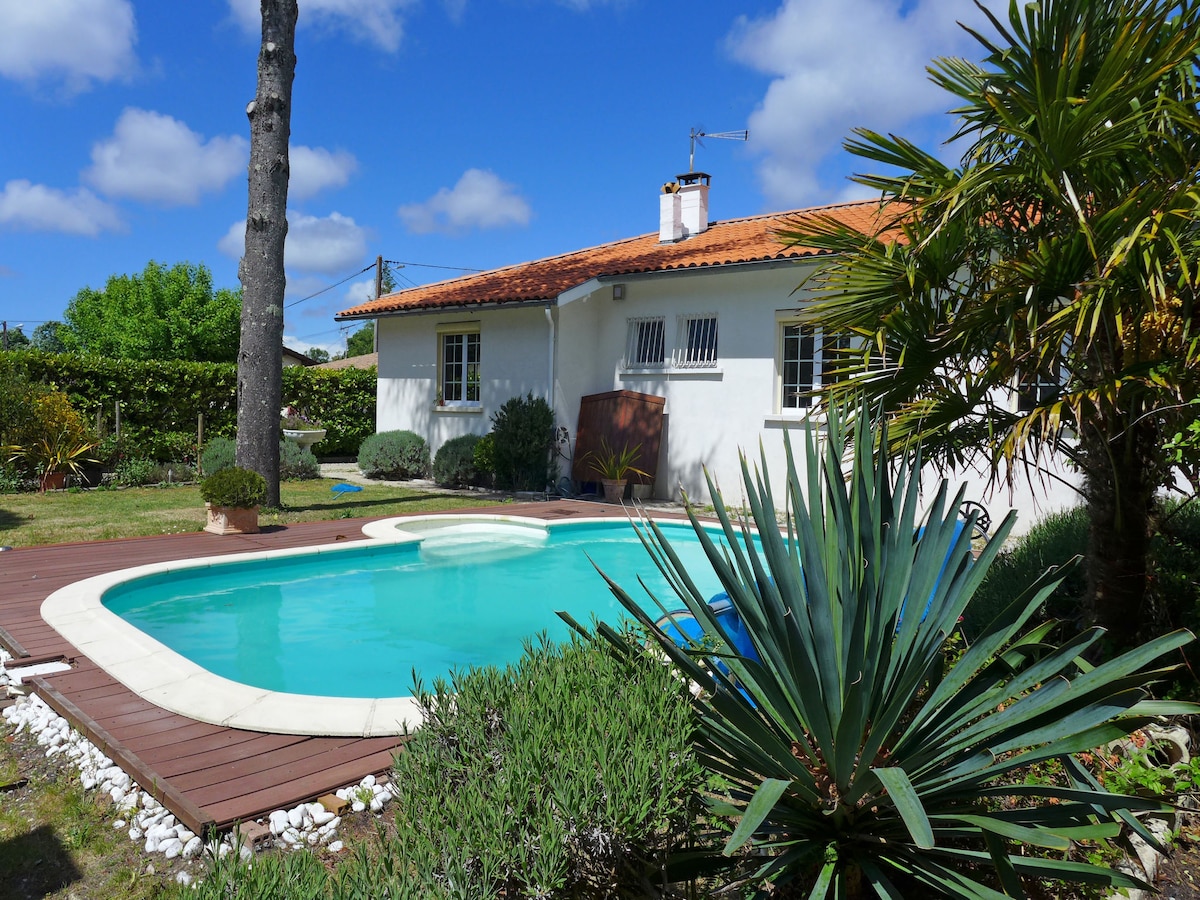 The property is surrounded by a well-maintained garden, featuring a seasonal swimming pool with a curved edge. The house displays a traditional style with a red-tiled roof and white exterior. Lush greenery and palm plants add to the outdoor ambiance, under a clear blue sky.