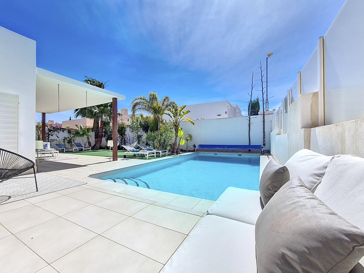 An inviting outdoor space is displayed, featuring a private swimming pool surrounded by sun loungers and palm trees. A shaded dining area is visible in the background, complemented by a clear blue sky. The contemporary architecture emphasizes relaxation and tranquility.