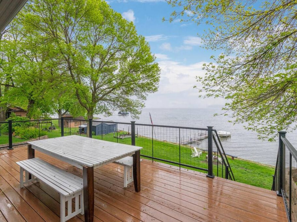A spacious wooden deck features a picnic table with benches, offering views of Lake Waconia. Lush green trees surround the area, and the calm water reflects the sky. A grassy slope leads down to the lake, enhancing the serene setting.