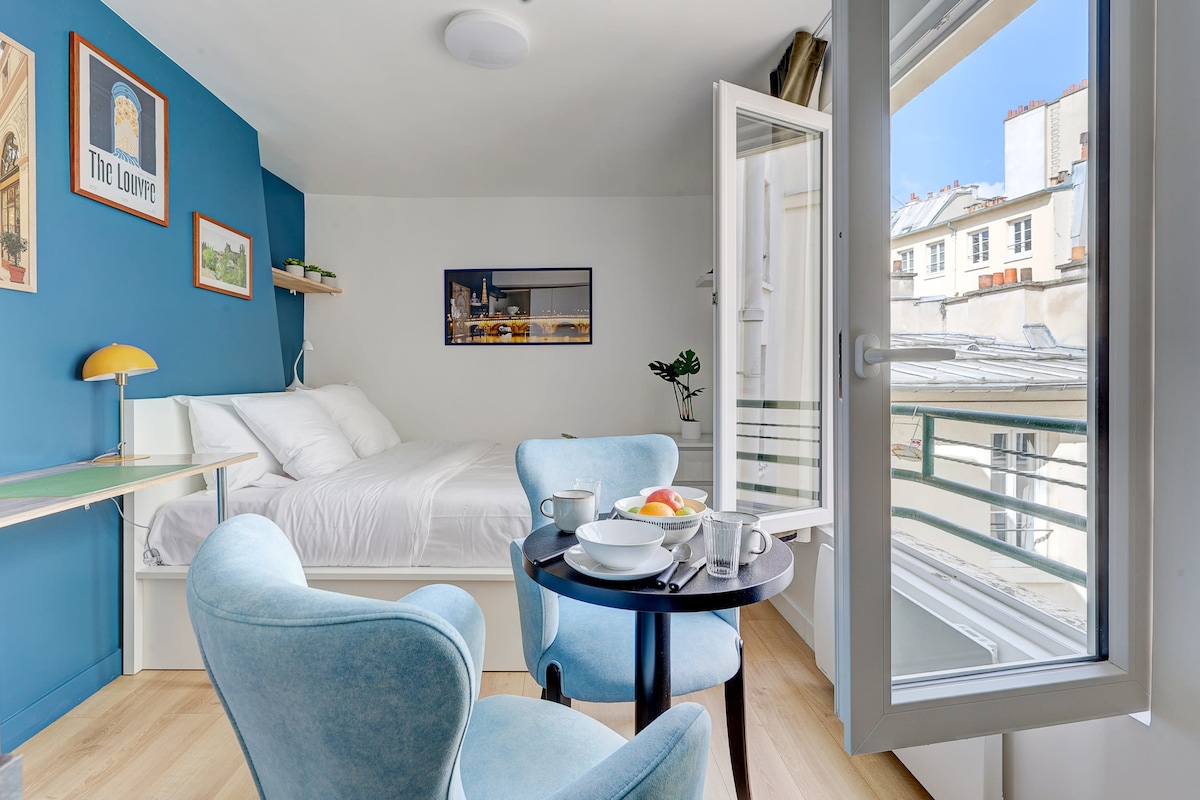 A bright studio apartment is showcased, featuring a cozy sleeping area with a spacious bed and blue accent walls. A small dining table is set with breakfast items, while large windows open to let in natural light and fresh air, enhancing the overall ambiance.