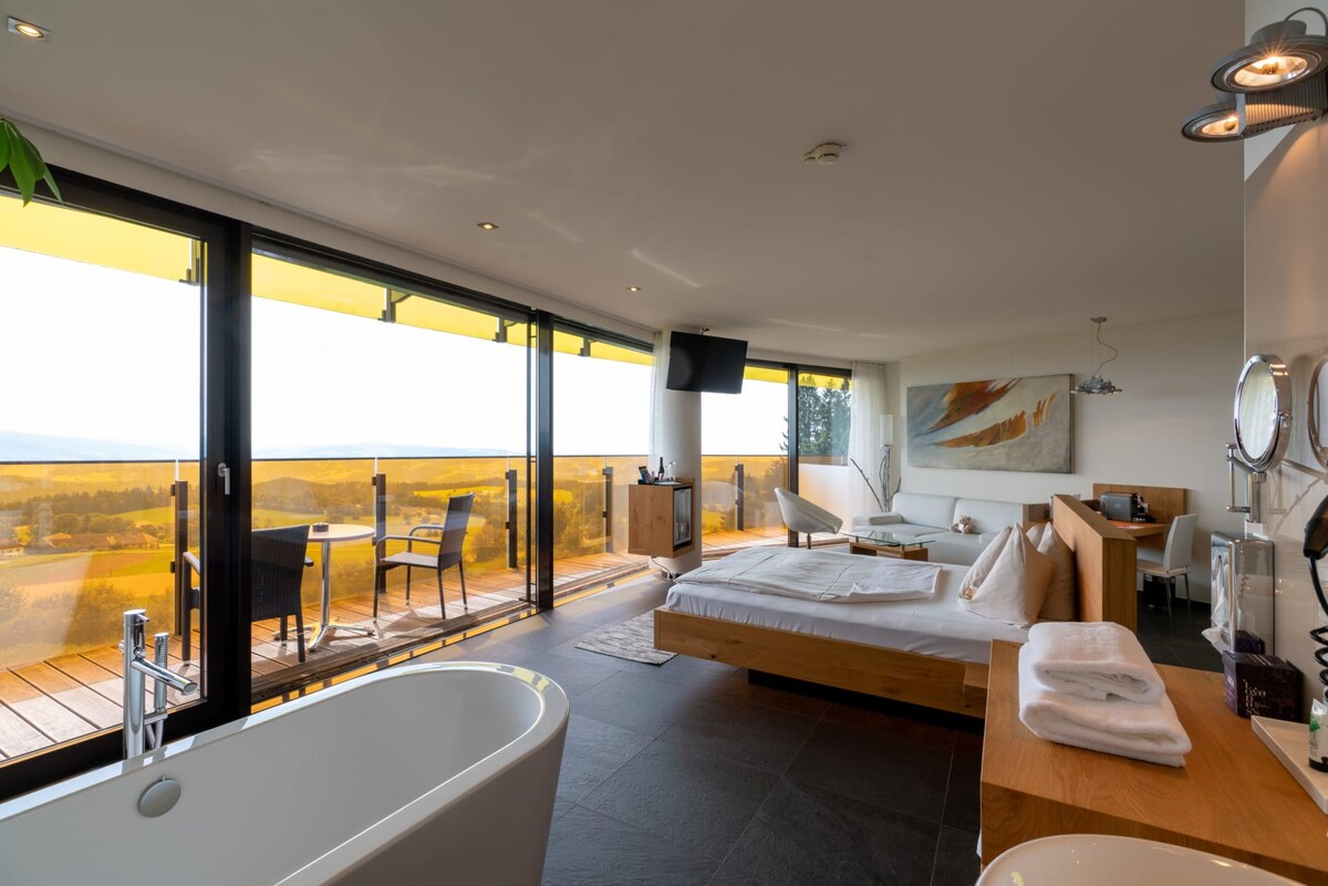 A bright and airy room features a spacious bed with crisp linens, alongside a freestanding bathtub. Large sliding glass doors open to a balcony with seating that offers expansive views of the surrounding landscape. Natural light floods the space, enhancing its serene ambiance.