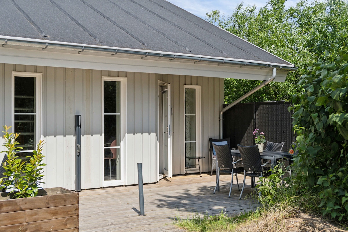 A private terrace area is presented featuring a wooden deck with six black chairs and a table, surrounded by green foliage. The entrance to the apartment is accessible from this space, with large windows allowing natural light to brighten the interior.
