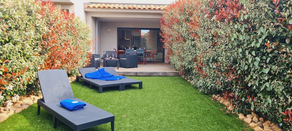 A well-maintained garden area features two lounge chairs with towels neatly placed on them. In the background, a terrace area with outdoor seating is visible, surrounded by vibrant green grass and colorful hedges, creating a serene outdoor space.