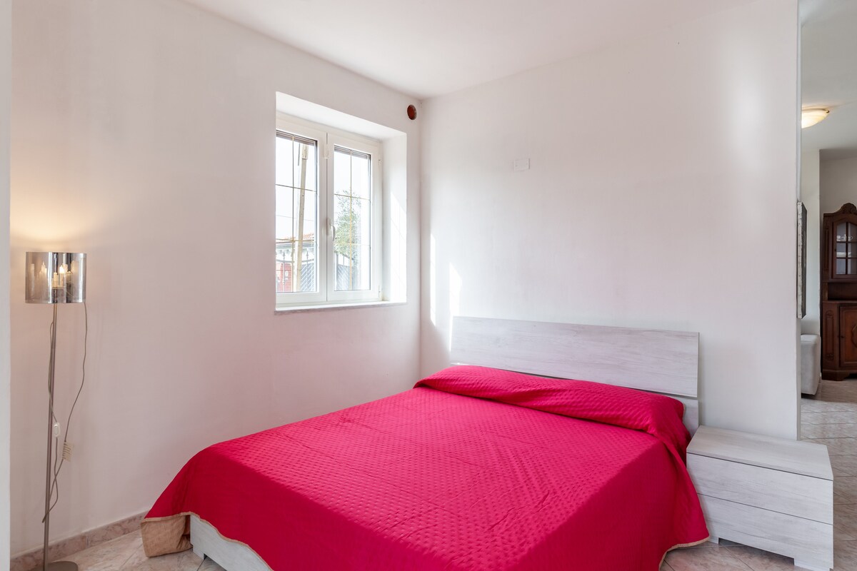A spacious sleeping area features a modern bed draped in a vibrant red coverlet, accompanied by a matching bedside table. Natural light enters through a large window, illuminating the clean, minimalist design of the room.