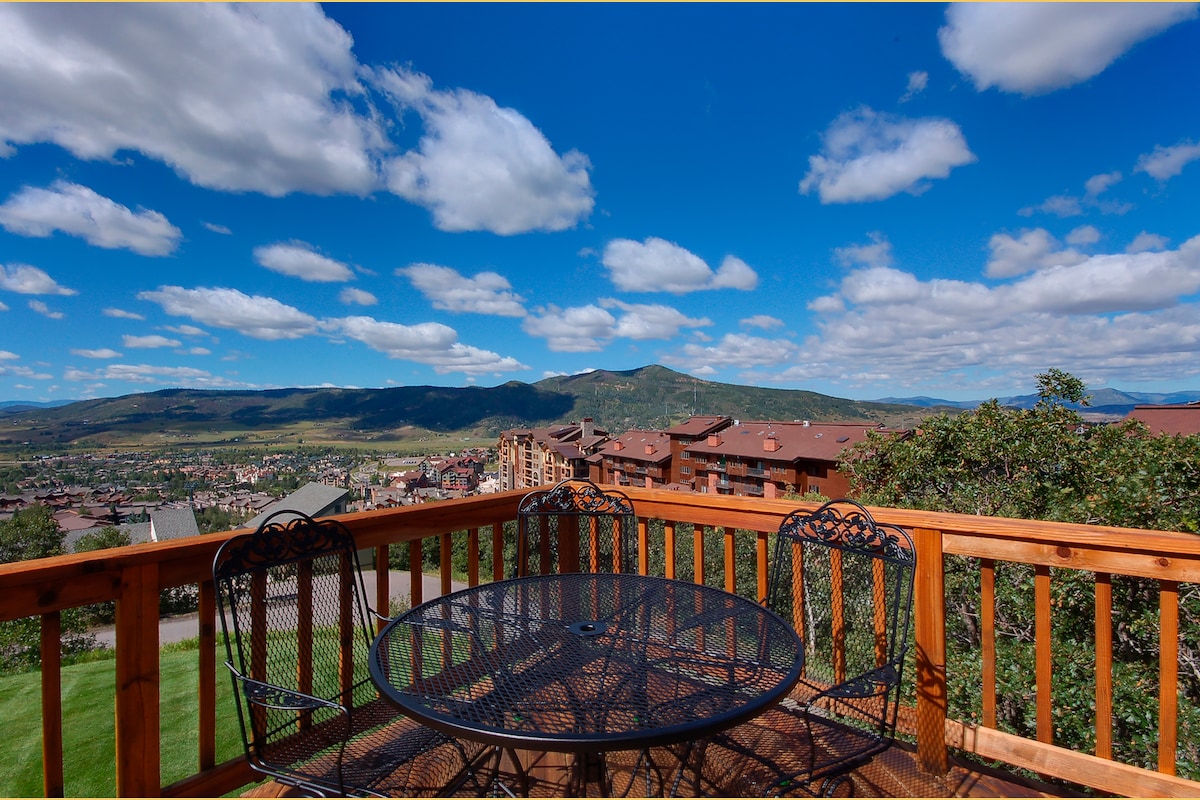 A spacious outdoor deck is featured, showcasing a round metal table surrounded by four matching chairs. The scenic view includes rolling mountains under a clear blue sky dotted with fluffy clouds, creating a serene backdrop for gatherings.