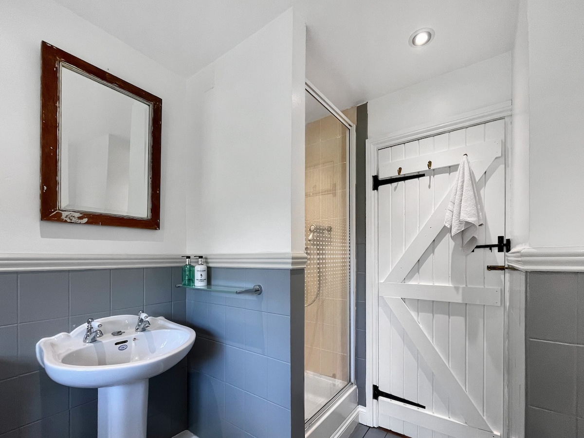 The bathroom features a spacious design, showcasing a porcelain sink with chrome faucets and a large mirror above it. A shower cubicle with glass doors is situated beside the entrance, accented by neutral tile tones and a white wooden door adorned with a towel hook.