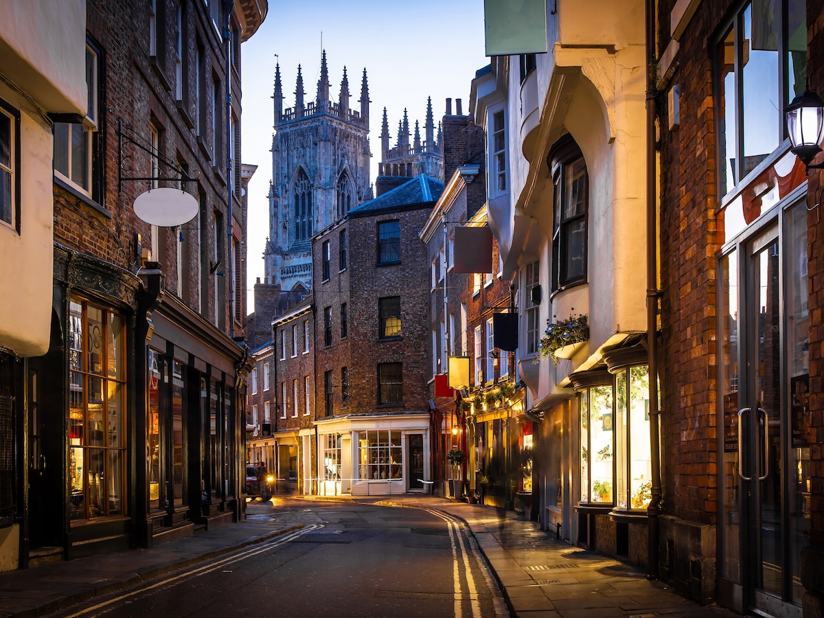 A charming street is lined with historic brick buildings, showcasing shopfronts with warm lighting. The iconic tower of a cathedral rises majestically in the background, framed by dusk's soft light. The cobblestone pathway invites exploration, highlighting the quaint atmosphere of this vibrant area.