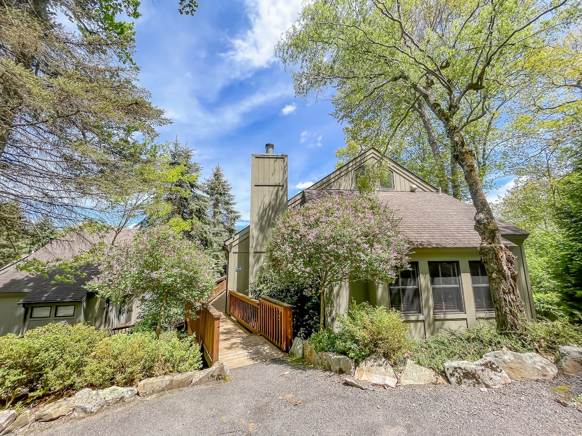 The exterior of the home is framed by lush greenery and blooming bushes, with a welcoming pathway leading to the entrance. A combination of wooden decking and stone accents enhance the natural surroundings under a clear blue sky, providing a serene first impression.