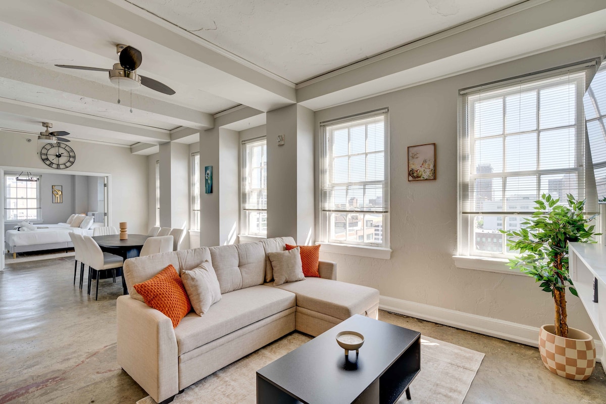 The spacious living room is filled with natural light from large windows, featuring a comfortable sectional sofa adorned with bright cushions. A sleek coffee table sits in front, complemented by a decorative plant and modern wall art, creating a welcoming ambiance.